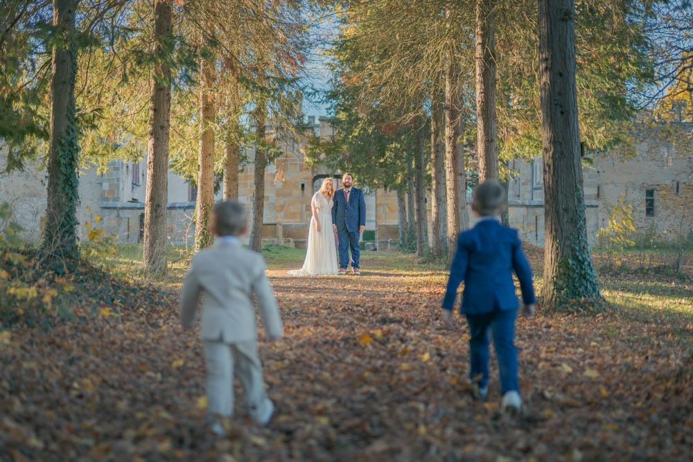 Couple marié debout dans une allée arborée d'un château regardant leurs deux enfants venir vers eux