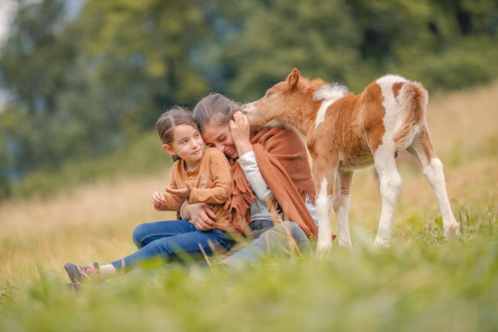 Une maman et sa fille riant d'un poulain venant mâchouiller les cheveux