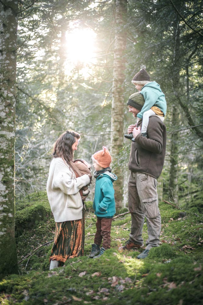 Photographie de famille en forêt, debout les pieds dans la mousse et la tête dans un rayon de soleil