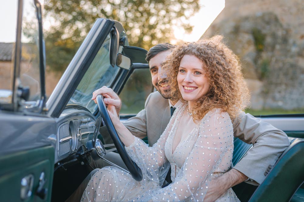 Couple de jeunes mariés assis dans leur voiture et souriant devant un château