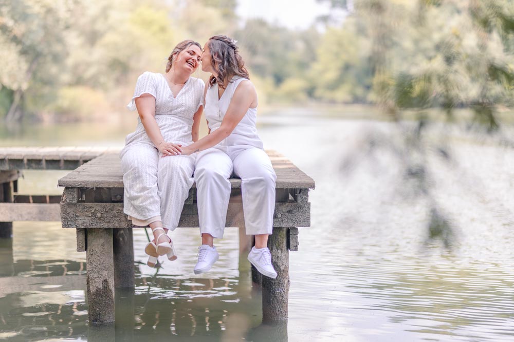 Couple assis sur un ponton à l'ombre des arbres se souriant l'un à l'autre
