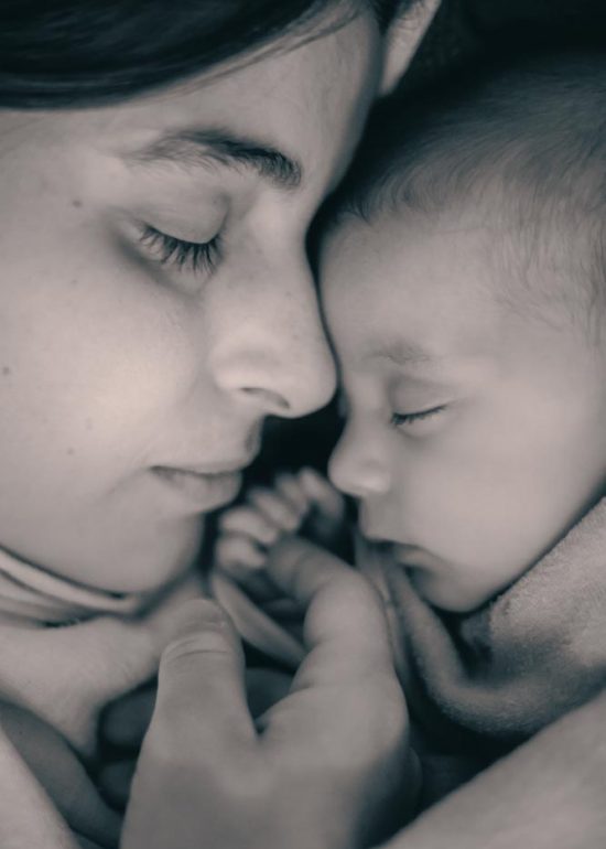 Une maman dort avec son bébé, collée à lui, leur front se touchant et la main du bébé tenant le bout d'un de ses doigts