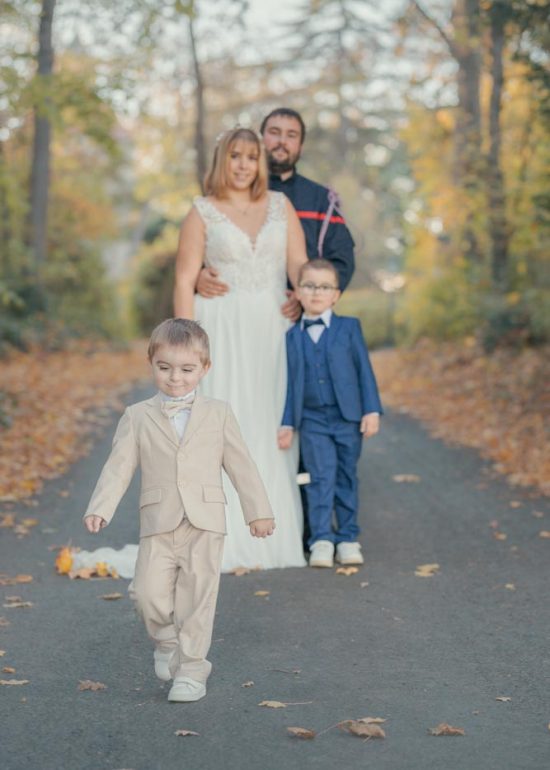 Un enfant s'éloigne souriant de ses parents, côté photographe, dans une allée arborée en automne alors que son frère reste avec les parents