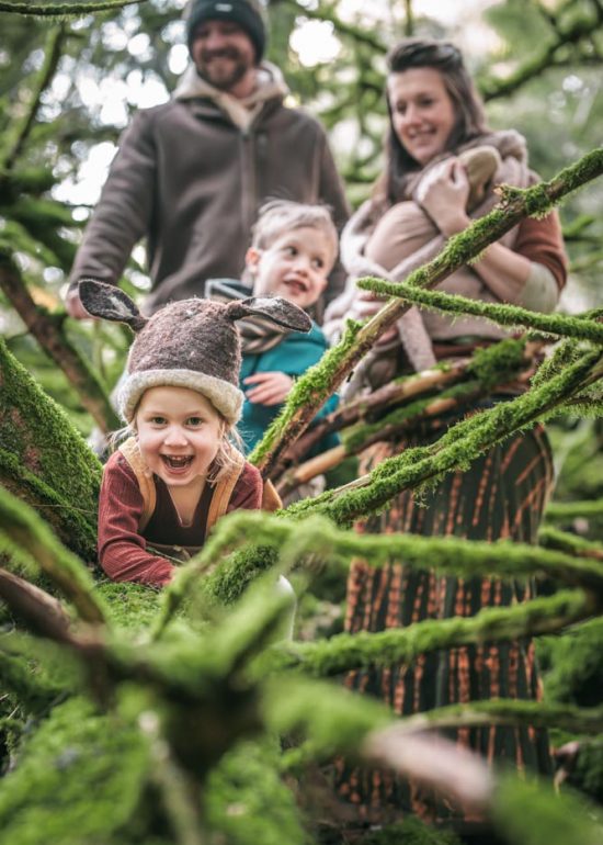 Famille de 5 enfants en forêt, deux enfants sont assis sur un tronc couché, leur fille la plus proche du photographe est même couchée sur le tronc en riant