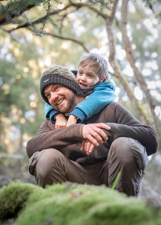 Portrait d'un papa et de son fils, le papa est accroupi en forêt et son fils est appuyé sur son dos, les bras ballant par dessus ses épaules