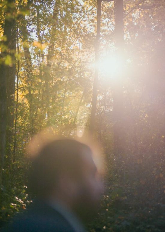 Plan sur la lumière dorée du soleil couchant à travers le feuillage d'automne d'une forêt, au premier plan flou on devine la tête d'un homme passant dans cette lumière