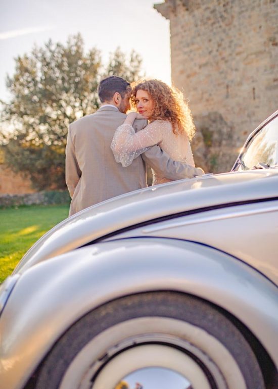 Couple de dos assis sur le capot d'une voiture de collection au coucher de soleil, la mariée regardant par dessus son épaule en direction du photographe