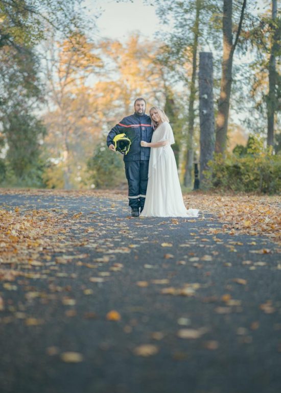 Couple de jeune marié posant dans une allée arborée couverte de feuilles tombées à l'automne, le mari est dans son costume de pompier