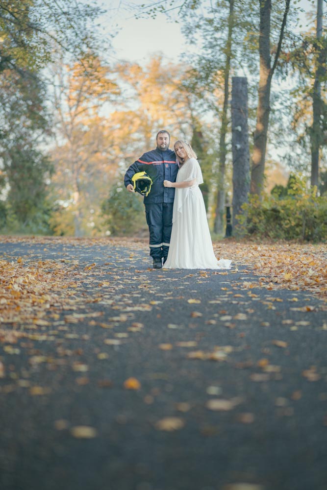 Couple de jeune marié posant dans une allée arborée couverte de feuilles tombées à l'automne, le mari est dans son costume de pompier