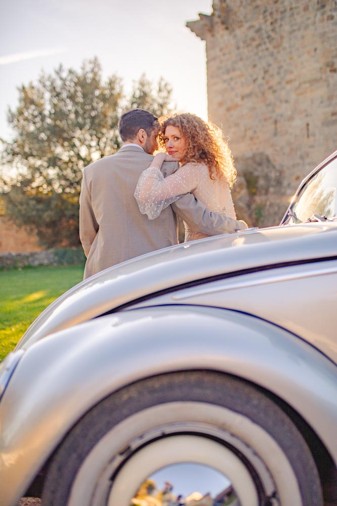Couple de dos assis sur le capot d'une voiture de collection au coucher de soleil, la mariée regardant par dessus son épaule en direction du photographe
