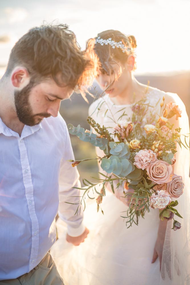 Couple de jeunes mariés, l'homme emmène sa femme qui porte son bouquet pastel, au coucher de soleil