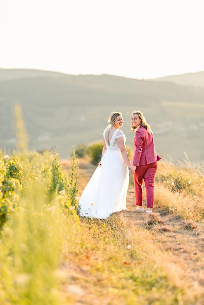 Couple s'éloignant dans un chemin bordant un champ de vignes, et toutes deux se retournent vers le photographe en souriant