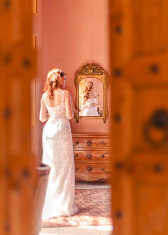 Photographie en plan large d'une mariée en pleins préparatifs, prise à travers la porte entrebâillée, son visage se reflétant dans un miroir