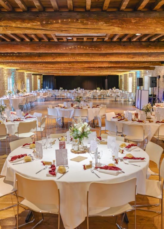 Plan large de la salle de réception d'un mariage, le bois apparent des poutres et du plafond donnant une ambiance ambrée à la salle baignée de lumière