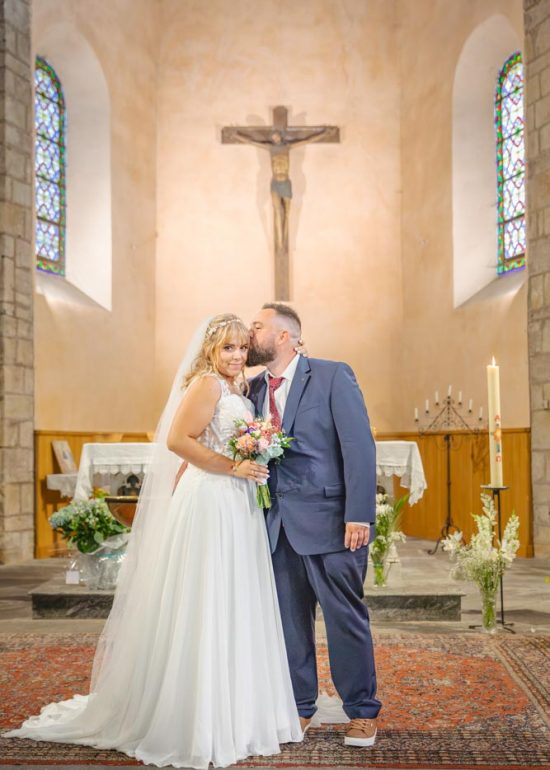 Photographie plein pied d'un couple enlacé dans l'église, le marié embrassant les cheveux de sa femme qui regarde le photographe