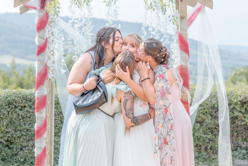 Photographie de groupe durant un mariage, les meilleures amies de la mariée l'embrassant toutes en même temps devant une arche de ruban et fleurs