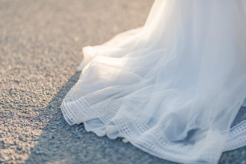 Détail d'une robe de mariée trainant sur le bitume d'une route au coucher de soleil