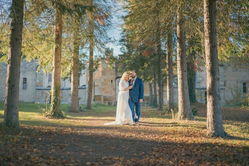 Couple posant debout l'un contre l'autre en plan large dans une allée arborée d'un château au soleil couchant à l'automne