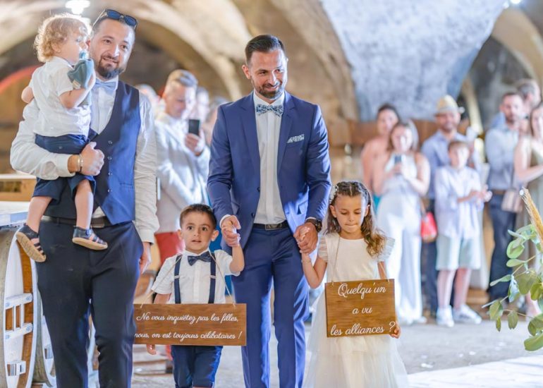 Trois enfants portant des pancarte lors d'une cérémonie disant "ne vous inquiétez pas les filles, nous, on est toujours célibataires" et "quelqu'un a vu les alliances ?"