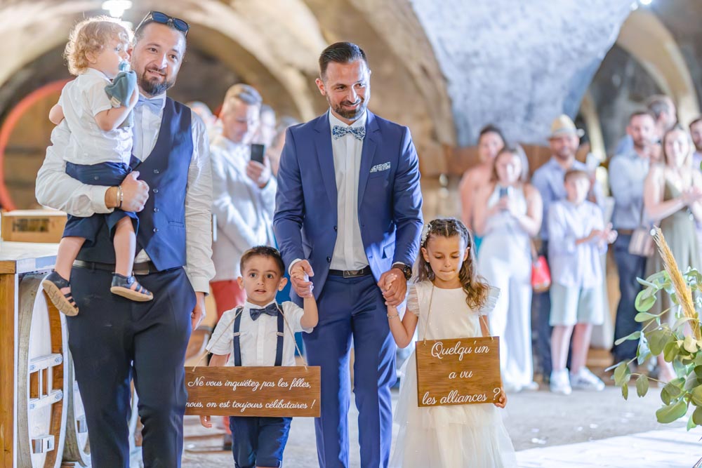 Trois enfants portant des pancarte lors d'une cérémonie disant "ne vous inquiétez pas les filles, nous, on est toujours célibataires" et "quelqu'un a vu les alliances ?"
