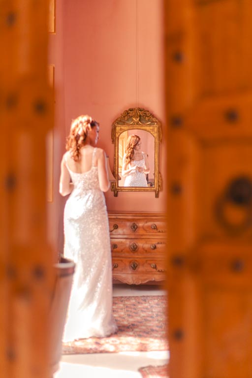 Mariée durant ses préparatifs prise en photo dans l'encadrure de la porte de la chambre et son visage se reflétant dans un miroir